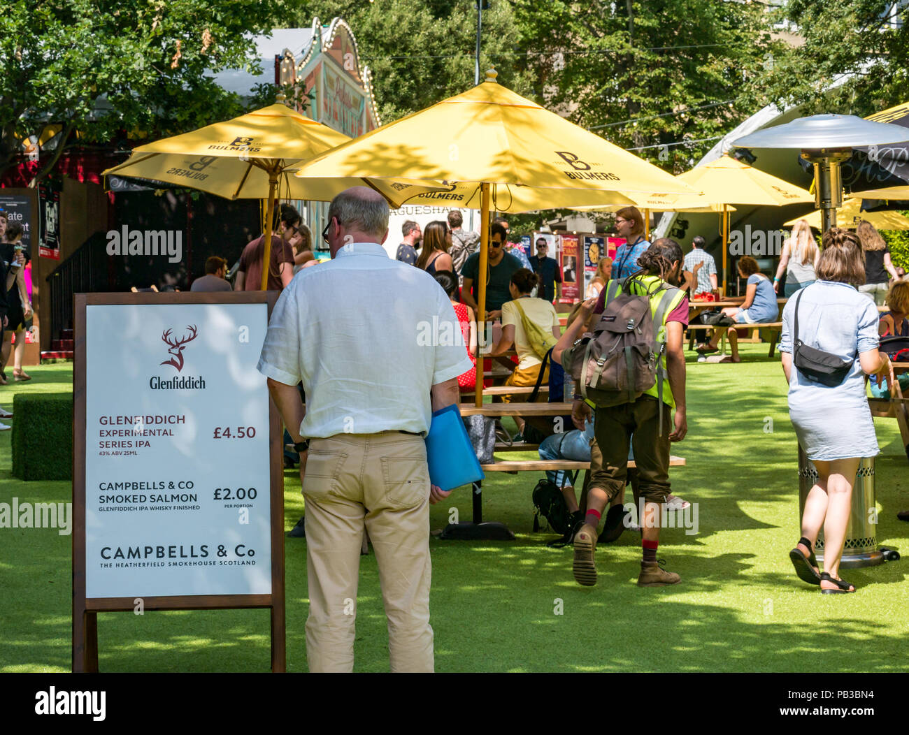 Edinburgh, UK. 26th July 2018. Edinburgh Food Festival 2018 in George Square Gardens, Edinburgh, Scotland, United Kingdom. There are food stalls  with over 20 local food and drink producers in the free outdoor event. People enjoying the sunshine in George Square at the food festival sitting at picnic tables under sun umbrellas. A man looks at the menu for Glenfiddich whisky and smoked salmon Stock Photo