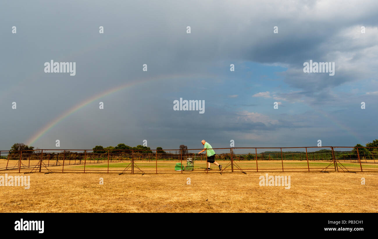Cricket pitch groundsman pushing a lawnmower underneath a rainbow and stormy sky Stock Photo