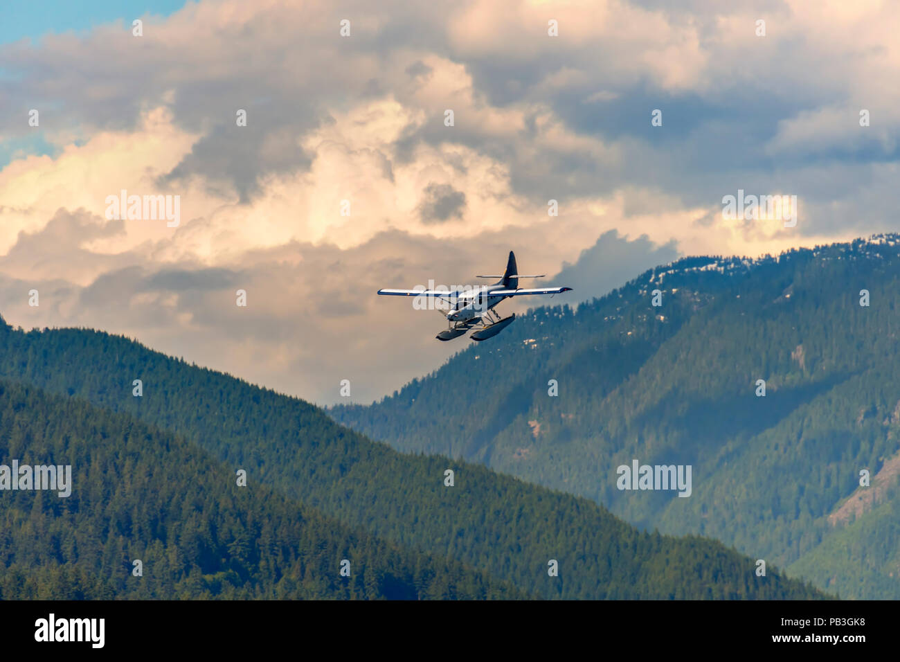 airplane, hydroplane flies in mountains covered with forest, under pink evening clouds and blue sky on a summer day Stock Photo