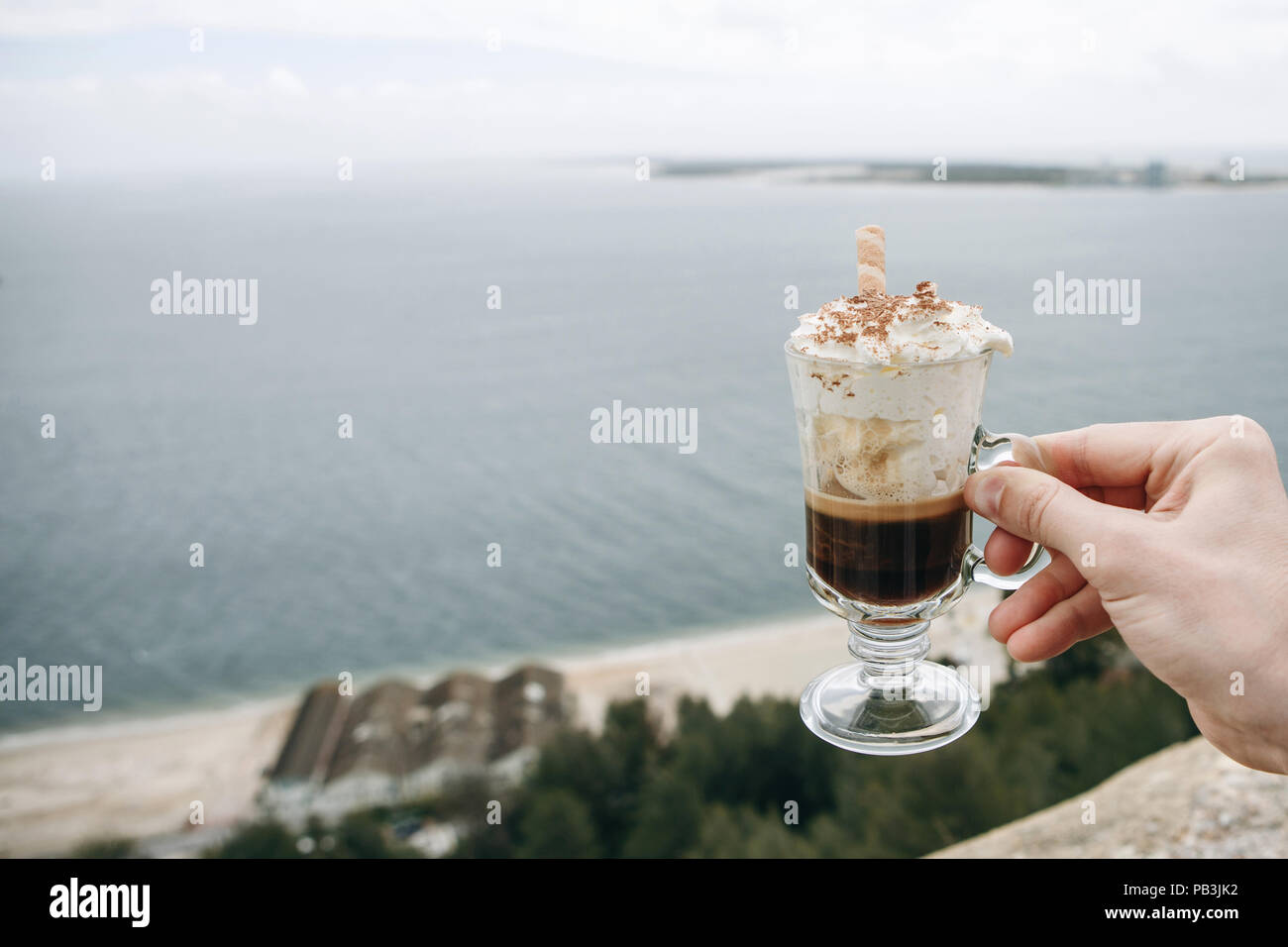 The man is holding a cup of fragrant Viennese coffee with whipped cream in the background of the Atlantic Ocean in Portugal and enjoying his rest. Stock Photo