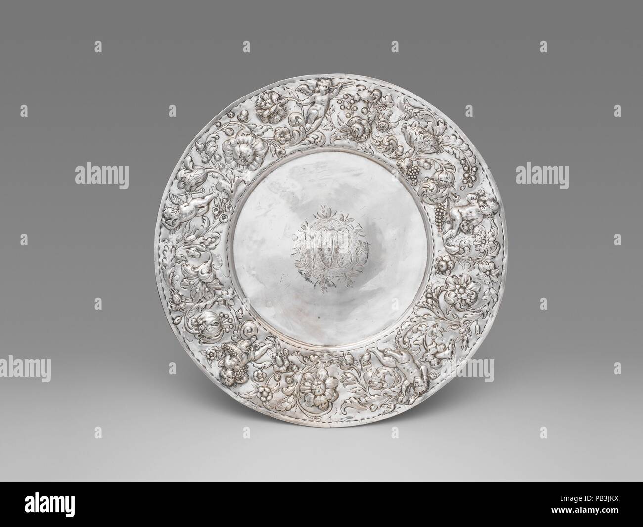 Dish. Culture: Dutch, Amsterdam. Dimensions: Diameter: 14 1/2 in. (36.8 cm). Maker: Sigismund Zschammer (ca. 1630, master 1661, d. before 1698). Date: 1685, with later monogram.  The cherubs, foliage and, particularly, flowers chased in high relief on the border of this dish were popular motifs not only in silver but also in contemporary painting and used for marquetry and gilt leather as can be seen in this and nearby galleries. The monogram engraved in the center refers to a member of De Peyster family, an important Dutch merchant family in New York which included Abraham de Peyster who serv Stock Photo
