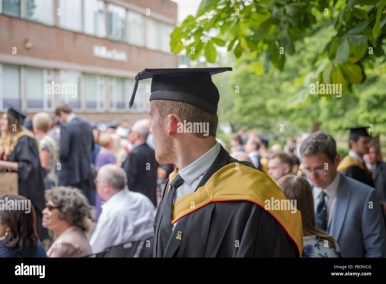 Young male graduated student seen his his graduation dress at a campus graduation party shown various parents in the background. Stock Photo