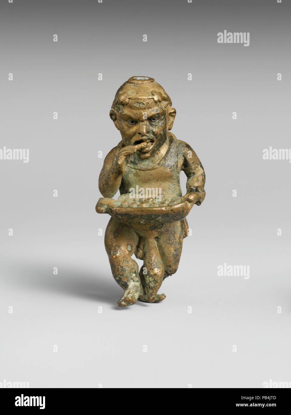 Bronze statuette of a dwarf with silver eyes. Culture: Greek or Roman. Dimensions: H. 3 1/8 in. (7.9 cm). Date: 1st century B.C.-1st century A.D..  The dwarf wears a wreath and carries a large tray of eatables from which he is helping himself. He may be a caricature of a street hawker at a festival. He has an apron tied at the back of his neck, and a bag hangs at his side. The type is Hellenistic, but the execution is probably Roman. Museum: Metropolitan Museum of Art, New York, USA. Stock Photo