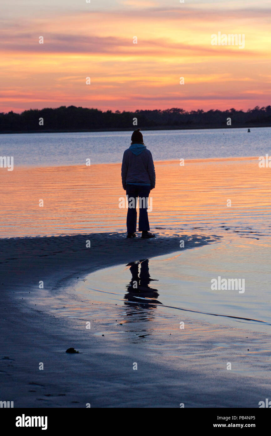 Silhouette of a woman standing alone at the water's edge of a beach at sunset enjoying nature and contemplating life Stock Photo