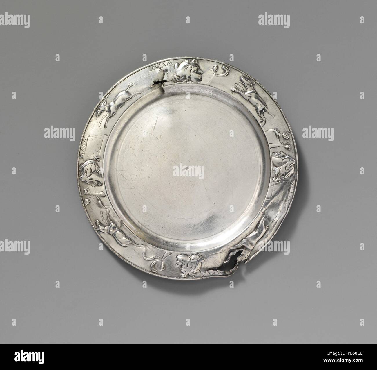 Silver plate. Culture: Roman. Dimensions: H. 1/16 in. (0.1 cm); diameter  5 in. (12.7 cm). Date: 1st-2nd century A.D..  The plate, originally part of a larger set of silver tableware, was clearly used over a prolonged period, since repeated polishing in antiquity has worn away the details of the rim decoration. On the underside are Latin inscriptions giving the names of two different owners. It was common for Romans to mark their valuable possessions in this way as a precaution against theft. The use of masks, symbols, and animals as decoration seen here was popular throughout the Imperial per Stock Photo