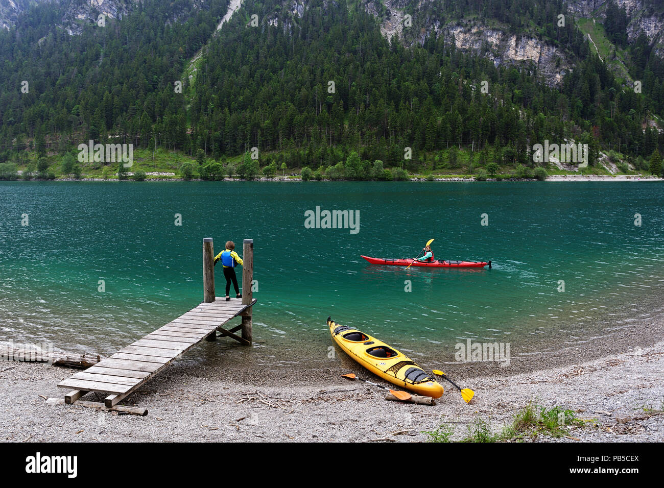 Mother in a kayak talking to her son standing on the wooden pier with a kayak parked on the shore on Plansee, Austria Stock Photo