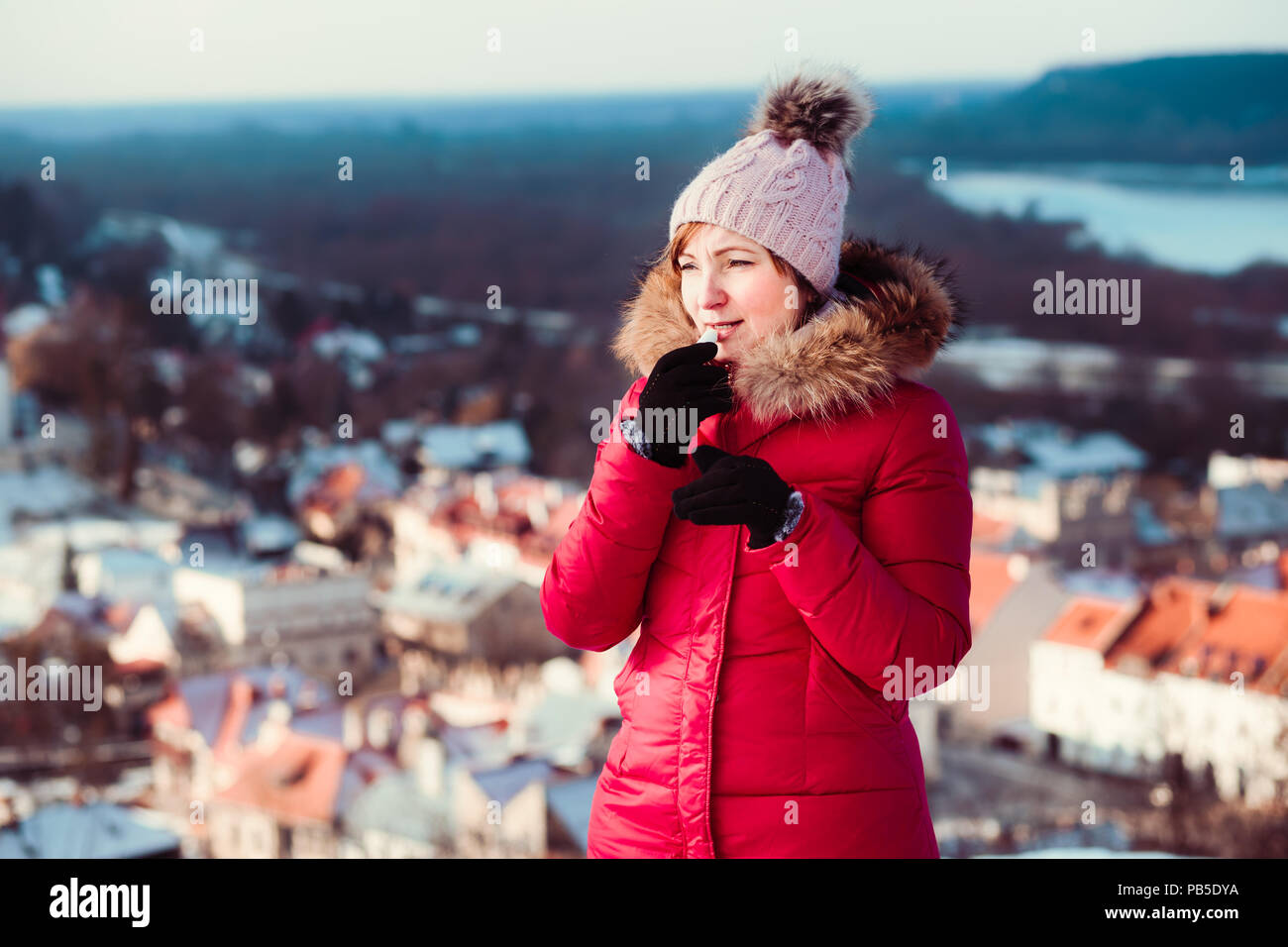 Woman applying lip balsam while walk in a wintery day. Wearing red coat and cap. Town in the background Stock Photo
