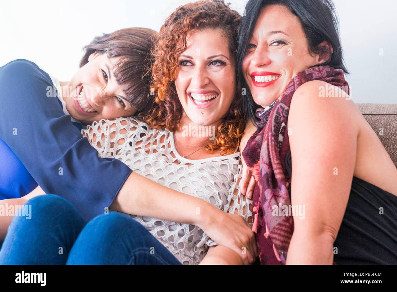 cheerful females middle age young womane friends together sitting on a sofa at home in leisure activity indoor. smile and enjoy the day hugging and to Stock Photo