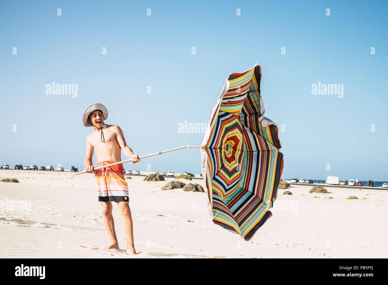 young male caucasian teenager play with umbrella sun and wind in a sandy paradise beach. smile and have fun enjoying the holiday and the warm weather Stock Photo