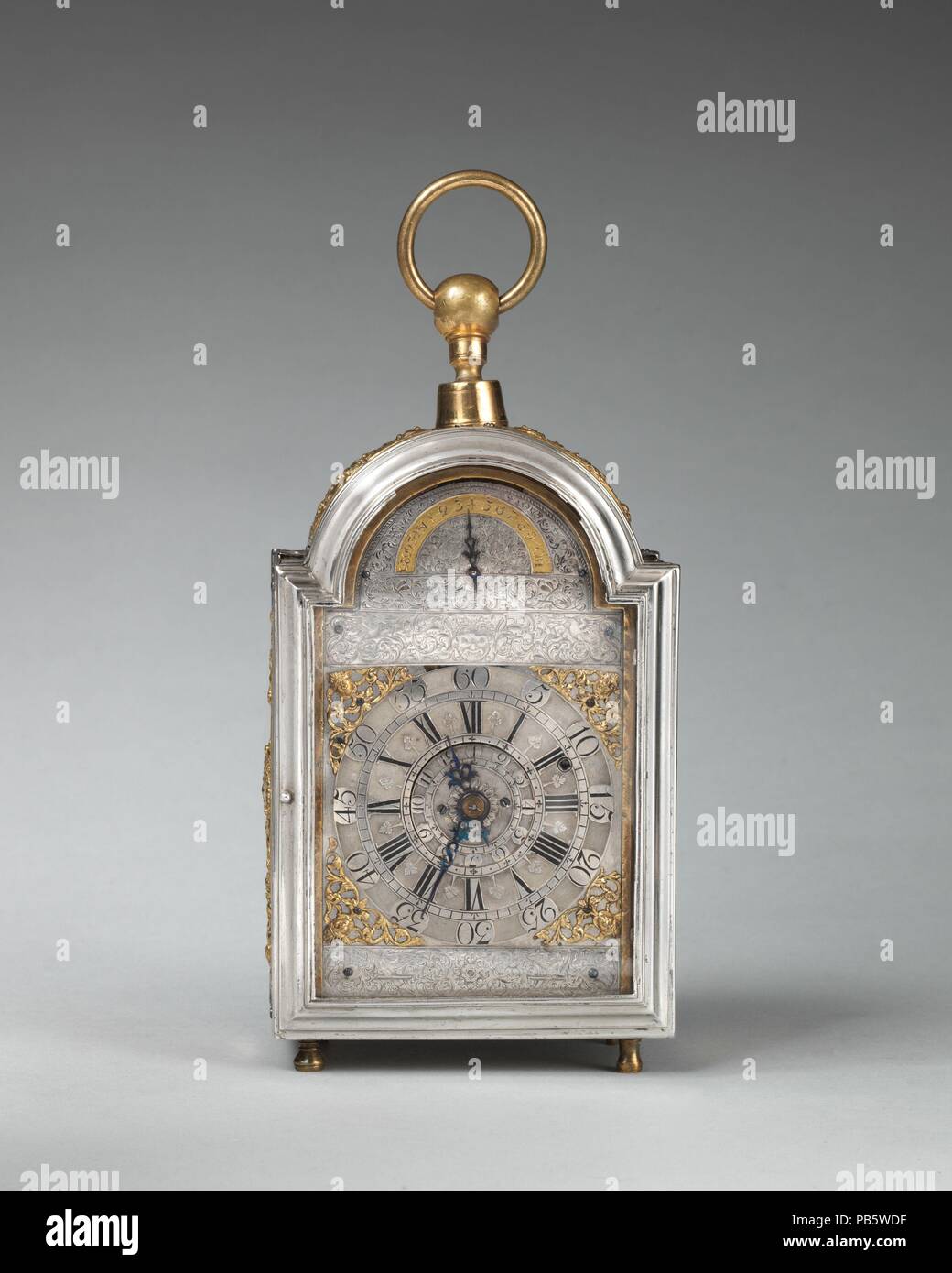 Traveling clock with alarm and calendar. Culture: British, London. Dimensions: Overall: 9 × 4 × 2 1/2 in. (22.9 × 10.2 × 6.4 cm). Maker: Probably by Joseph Paulet (recorded 1687-1716). Date: ca. 1700-1710.  In the late seventeenth and early eighteenth century small clocks adapted for traveling were comparatively rare. This example, in an exquisite case of engraved and repoussé silver, with a champlevé silver dial, belongs to a group of five or six examples, all signed 'Paulet London.' The universal ball joint at the top allows the clock to swing from its suspension ring, cushioning the jolts o Stock Photo