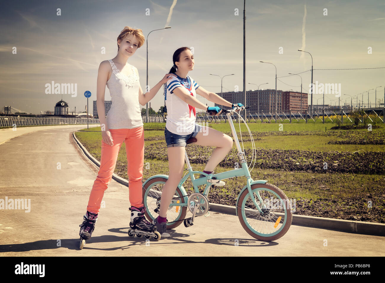 Two young female friends one on a bicycle, one on a roler scates. Summer sport and leisure theme. Warm toned image Stock Photo