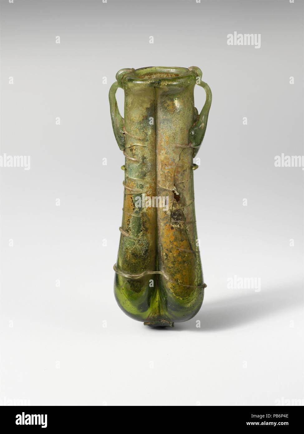 Glass double cosmetic flask (kohl tube). Culture: Roman, Syrian. Dimensions: H.: 4 3/8 x 1 3/4 in. (11.1 x 4.4 cm). Date: 4th century A.D..  Translucent yellowish green; handles and trail in same color.  Tubular rim folded out, over, and in; body comprising two tubes, side by side, made from a single inflated gather by pinching sides vertically to make a diaphragm; thick, flattened bottom with prominent pontil pad; two rod handles attached to side of body over trail in large, claw pads, drawn up and out, turned in, and trailed off on edge and top of rim.  Single trail applied as a pad on upper Stock Photo