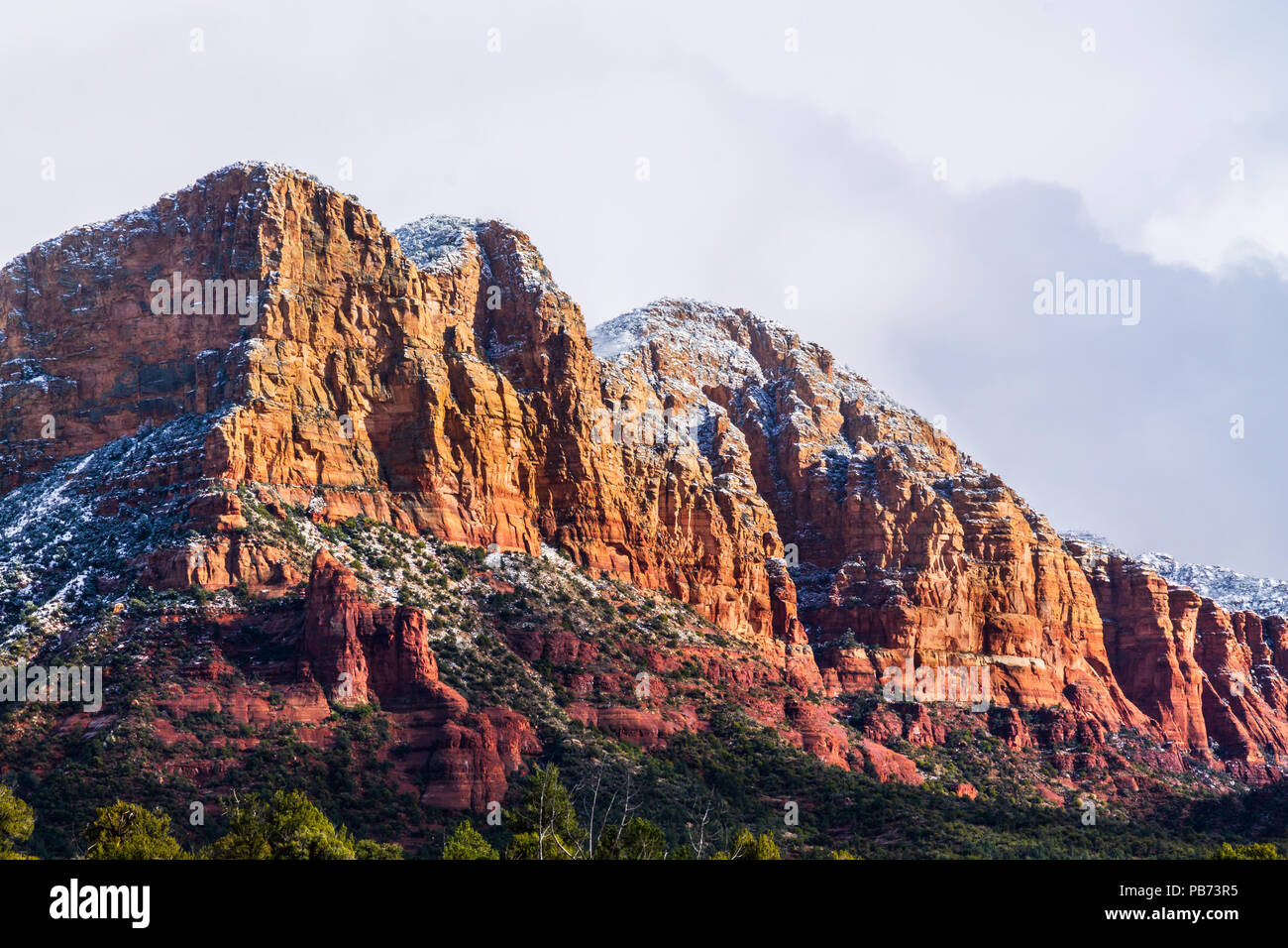 Rocky, rugged red sandstone hillside in Sedona, Arizona. Top is sprinkled with snow, cloudy sky is in the background. Sun is shining on one side. Stock Photo