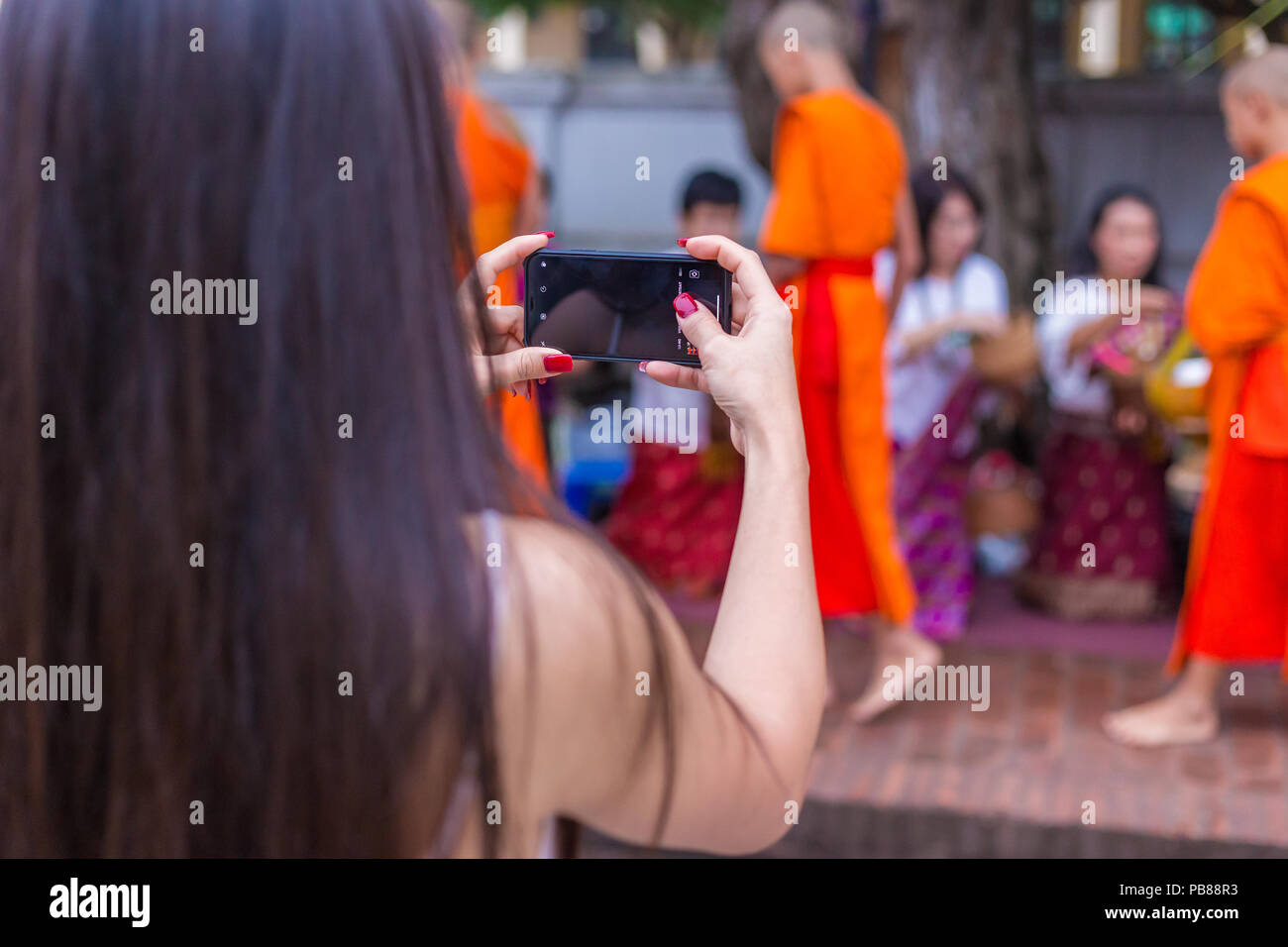 LUANG PRABANG, LAOS - 29 JUNE 2018 - A female tourist takes picture of people in traditional clothes giving food to buddhist monks in the morning of J Stock Photo