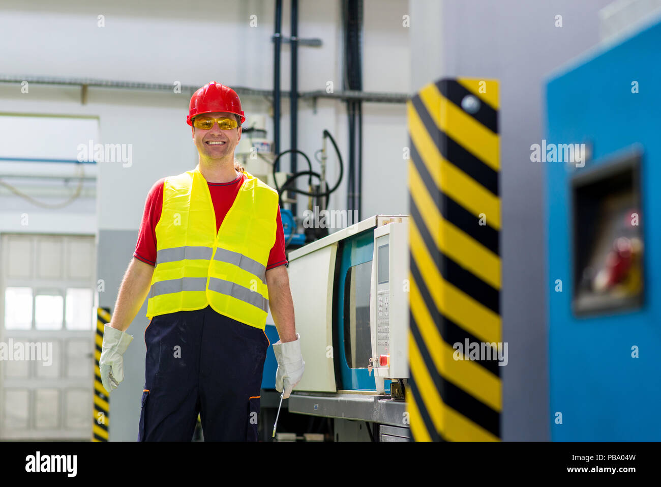 Factory worker smiling and looking at a camera standing beside a factory machine. Worker wearing safety clothing. Stock Photo
