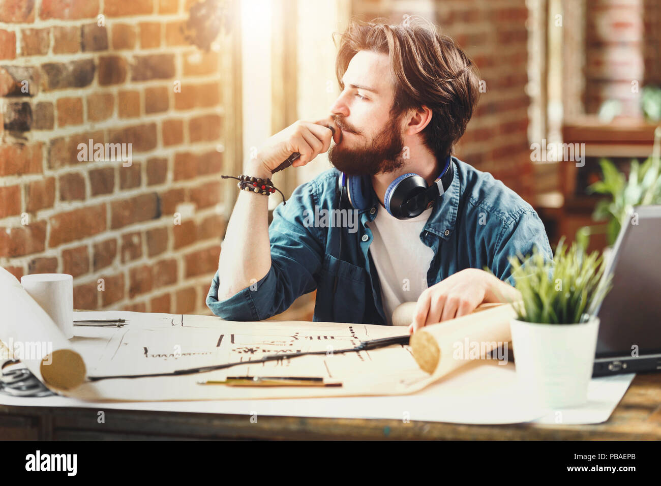 Concentrated young architect lookingto the window and thinking about new blueprint while sitting with laptop in a loft sunny office Stock Photo