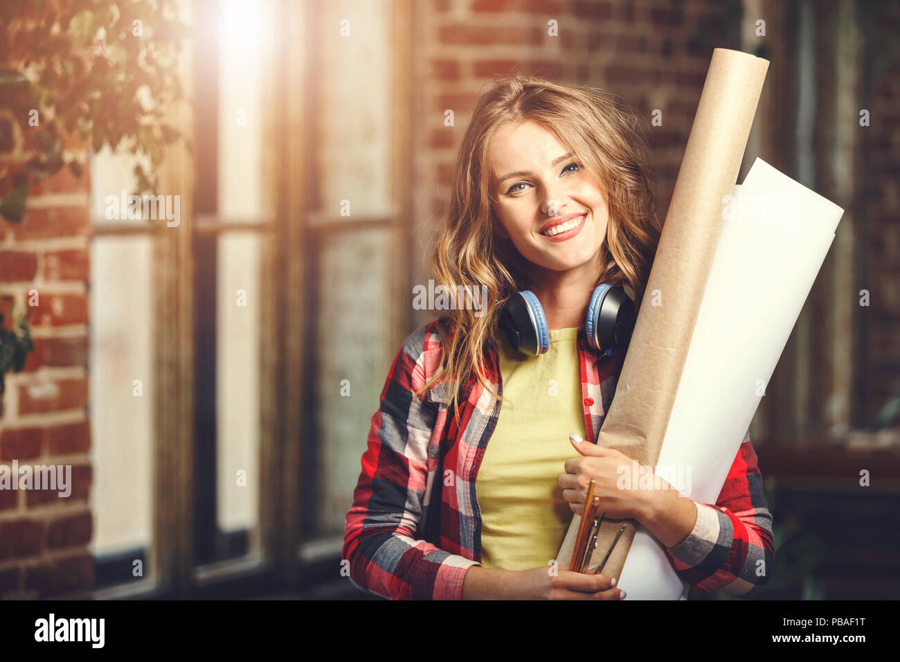 Handsome young long-hair female architect with headphones holding blueprints and smiling near the window in the loft modern office Stock Photo
