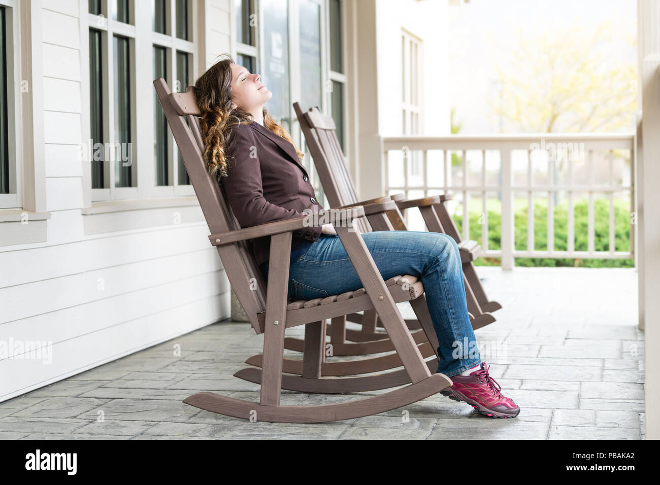 Young woman sitting on reclining, rocking chair, recliner, rocker by house, home, townhouse with closed eyes, enjoying fresh air, nature, sunset outsi Stock Photo