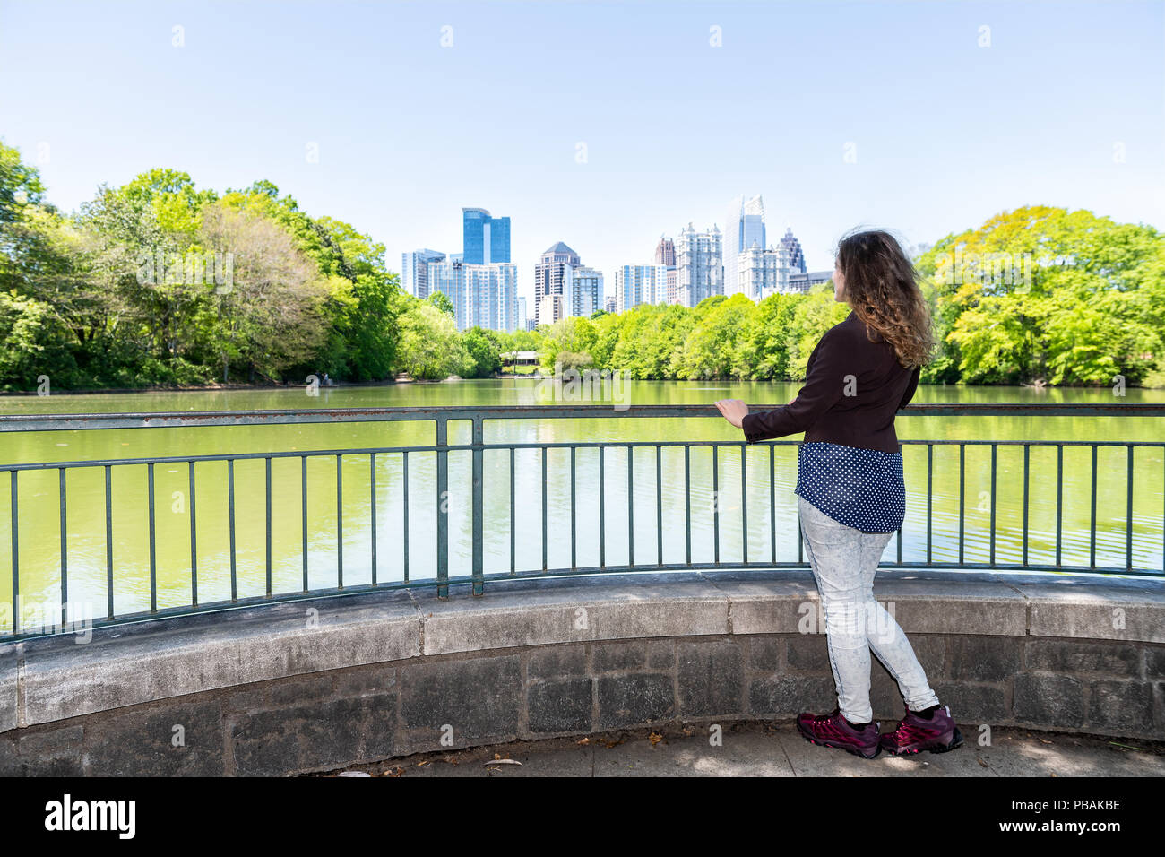 One young woman standing in Piedmont Park in Atlanta, Georgia looking at scenic view, water, cityscape, skyline of urban city skyscrapers downtown, La Stock Photo