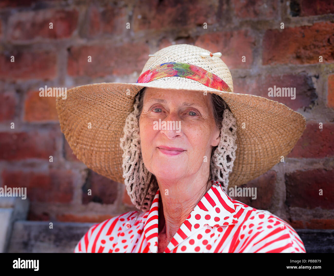Close, front view portrait of isolated senior woman, hair in crocheted snood wearing wide-brimmed floppy straw hat, 1940's summer event, UK. Stock Photo