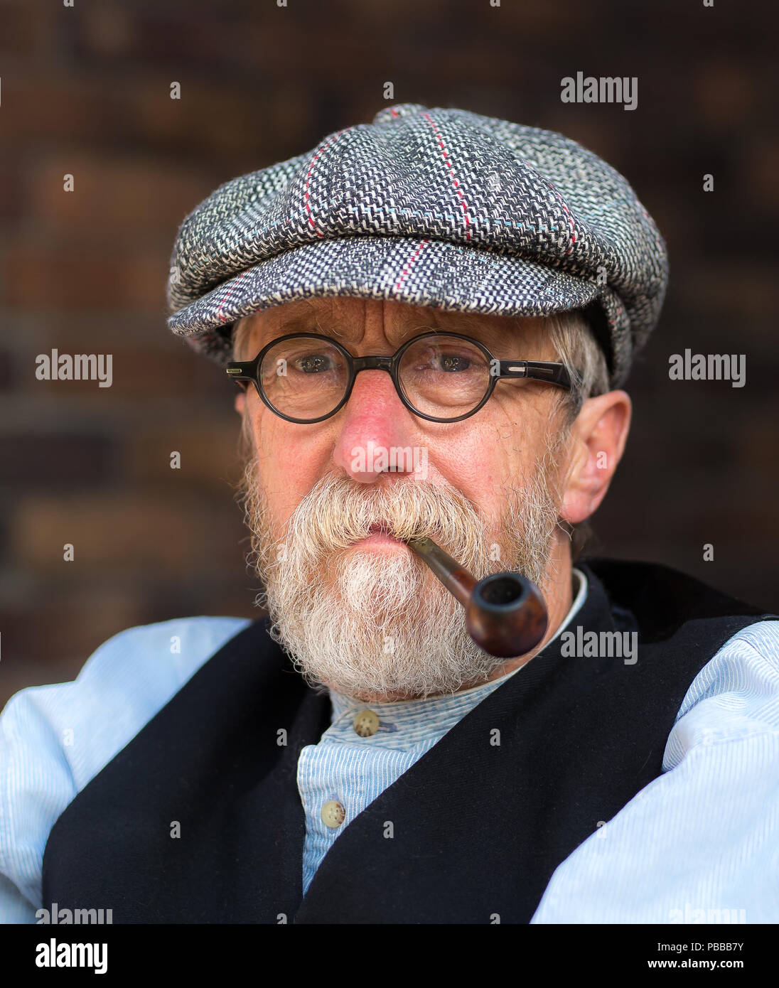 Close-up, front view portrait of senior man, bespectacled with pipe in mouth, wearing flat cap at Black Country Living Museum WWII 1940's event. Stock Photo