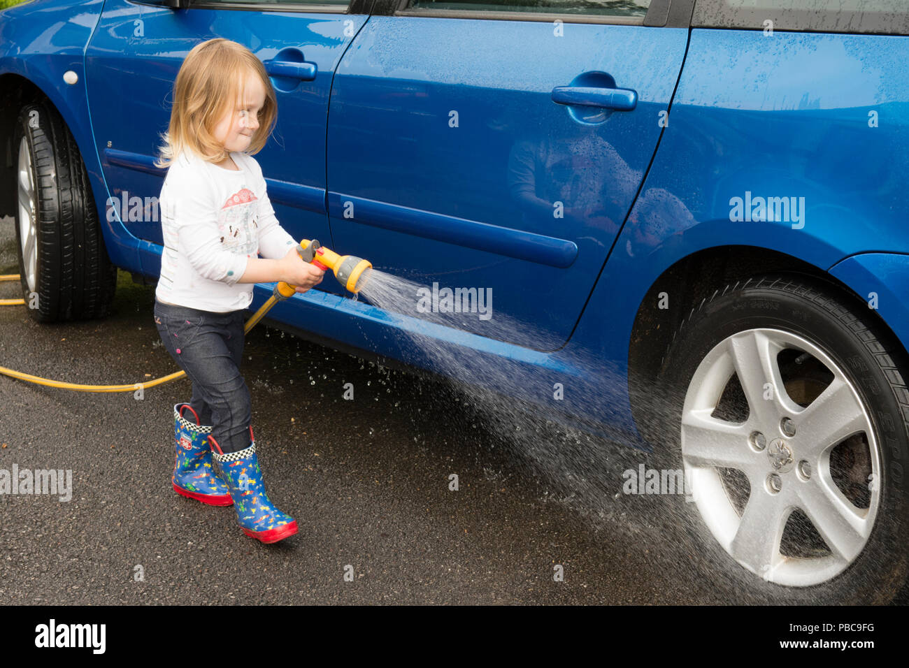 Three year old girl holding using hose pipe helping to wash blue car, spraying bodywork, UK. Stock Photo
