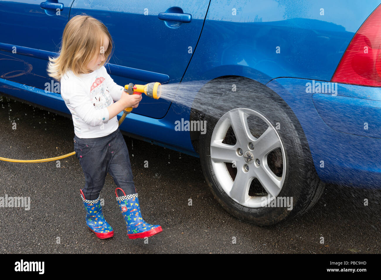 Three year old girl holding using hose pipe helping to wash blue car, spraying bodywork, UK. Stock Photo