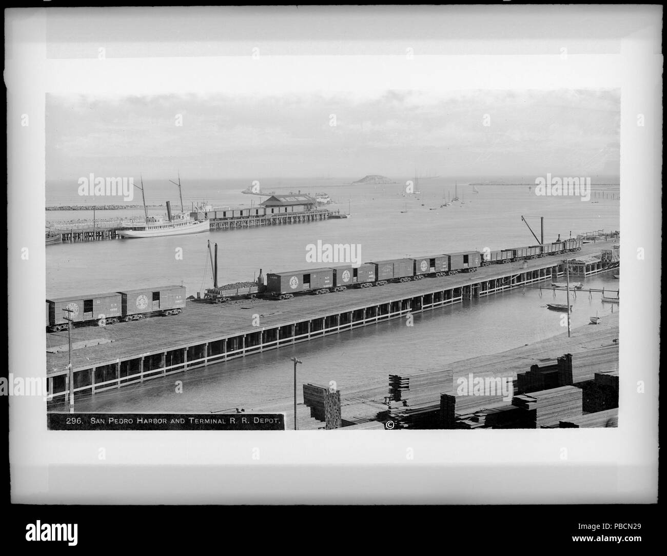 . English: Port of Los Angeles, unloading lumber, and railroad cars Photograph of the Port of Los Angeles, San Pedro Harbor and Terminal Railroad Depot, around 1905. The view is from the dock southwest to the mouth of the breakwater. Deadman's Island has not yet been removed, it is visible on the left side of the mouth. The ships which appear include both steam and sailing vessels. A railroad runs in the foreground, to the docks on which lumber has been unloaded. Another railroad appears in the background, on the other side of the channel. As Los Angeles tripled its population between 1900 and Stock Photo