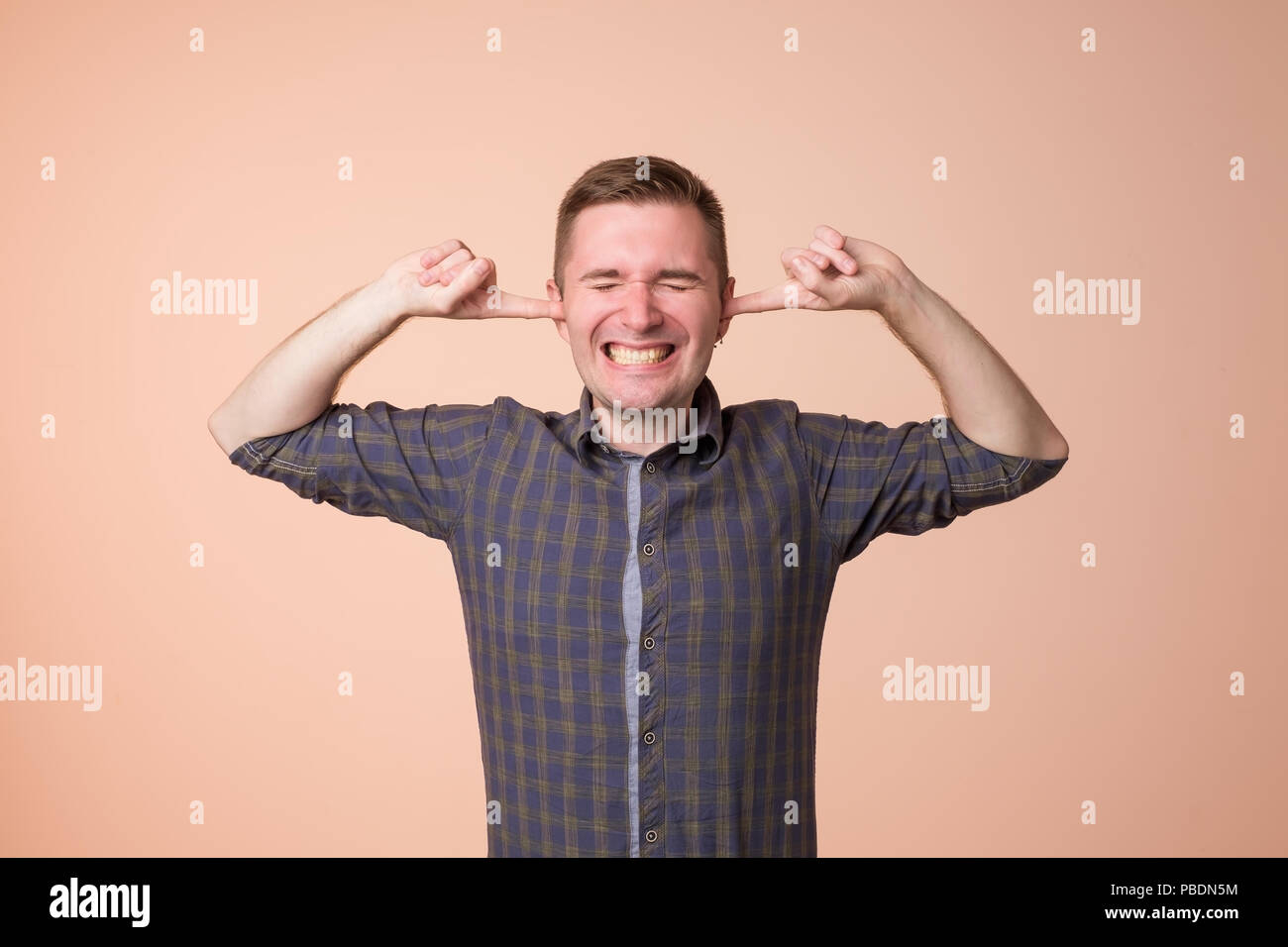 Studio portrait of young, unhappy, stressed man covering his ears, standing on dark gray wall background. Stock Photo