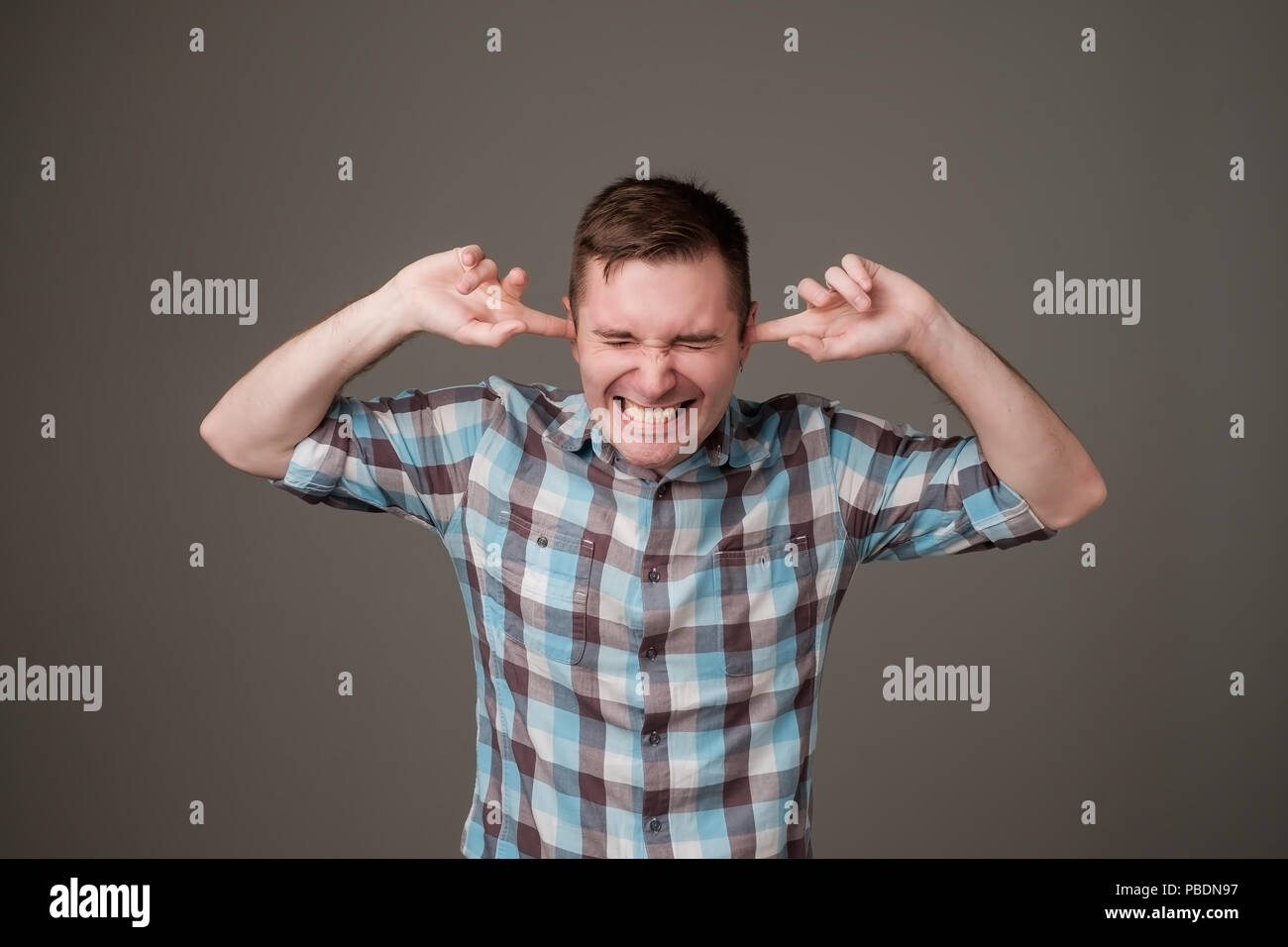 Studio portrait of young, unhappy, stressed man covering his ears, standing on dark gray wall background. Stock Photo