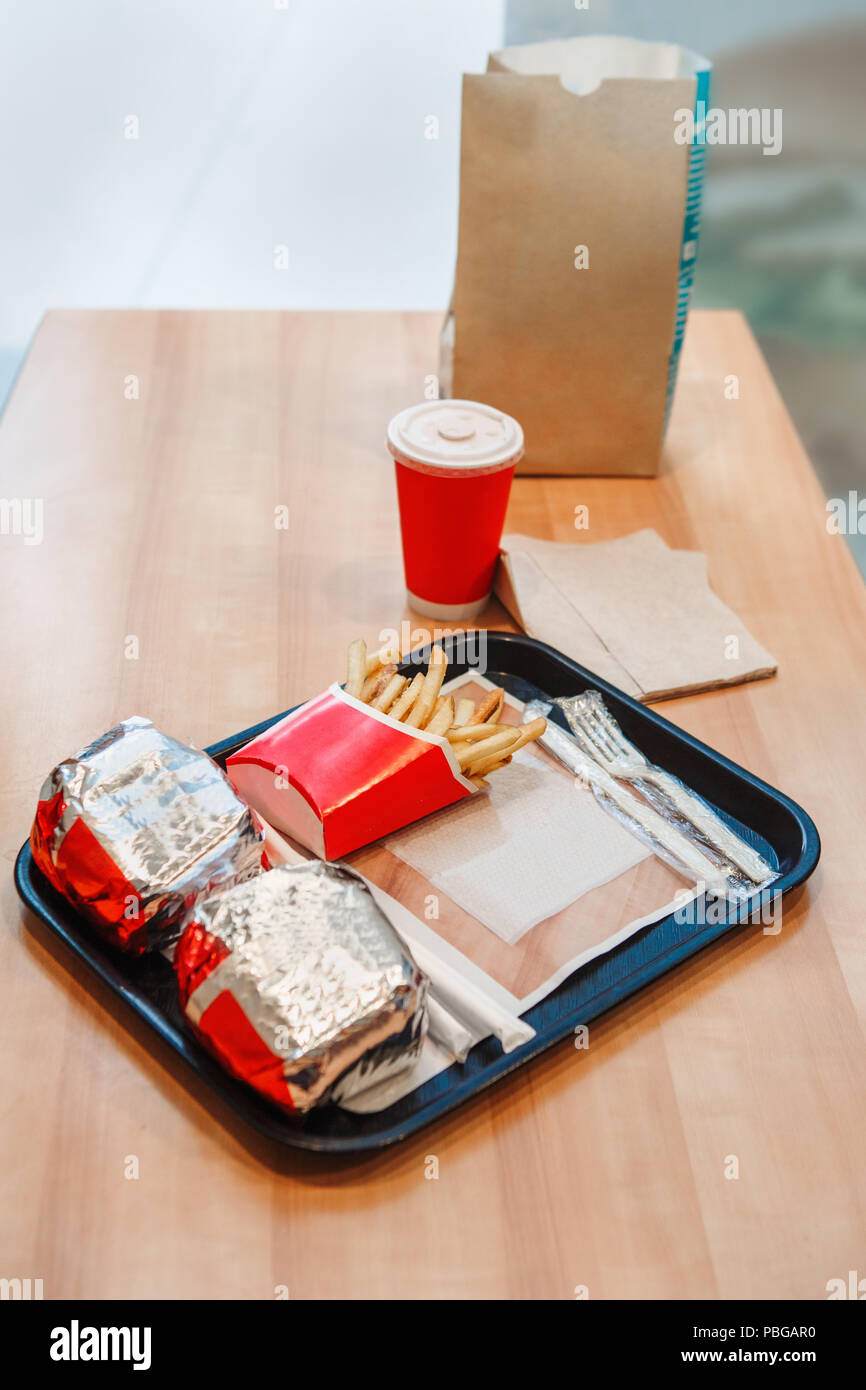 Closeup shot image of two burgers wrapped in tinfoil, french fries potato chips, red cup of coffee or tea on tray on wooden table in fast food restaur Stock Photo