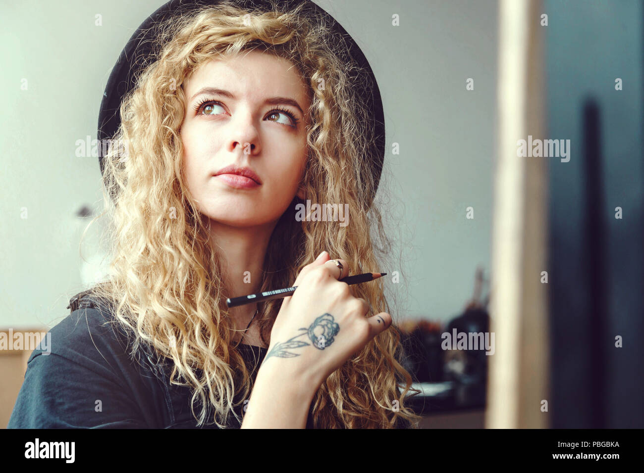 Portrait of long hair curly girl in handsome black hat sitting and dreaming with pencil behind the easel in studio Stock Photo