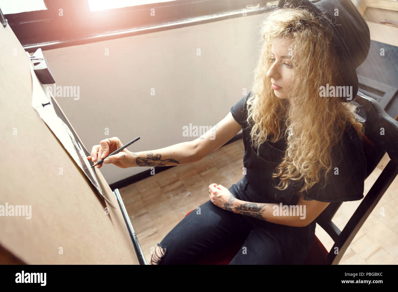Long hair curly girl in cute black hat drawing sketch behind the easel in sunny studio Stock Photo