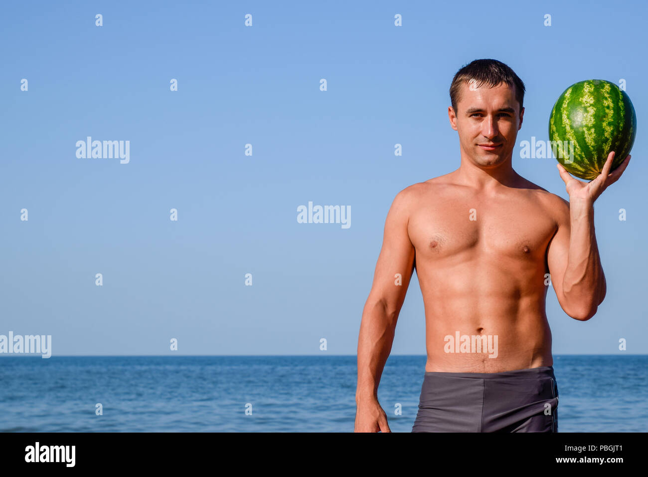 A sporty man is standing on the beach and holding a whole watermelon. A ripe watermelon in the hands of a man. Stock Photo