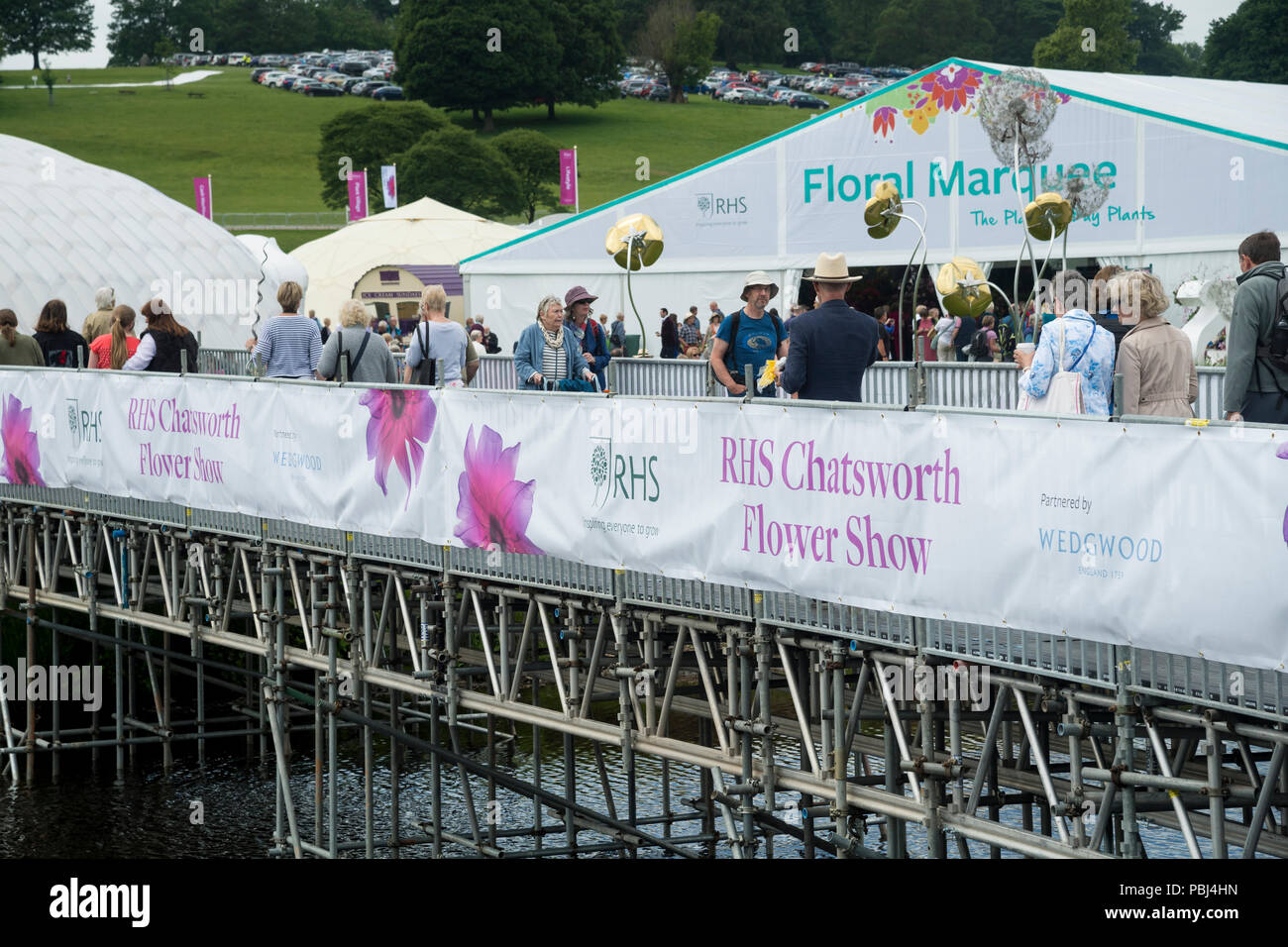 People coming & going at busy RHS Chatsworth Flower Show, some walking & crossing temporary river bridge by large marquees - Derbyshire, England, UK. Stock Photo