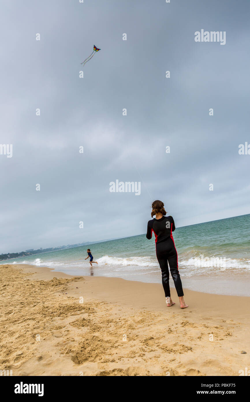 Two happy kids flying a kite on sandy beach wearing wetsuits Stock Photo