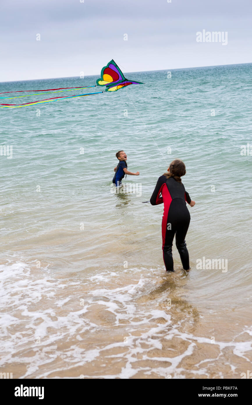 Two happy kids flying a kite on sandy beach wearing wetsuits Stock Photo