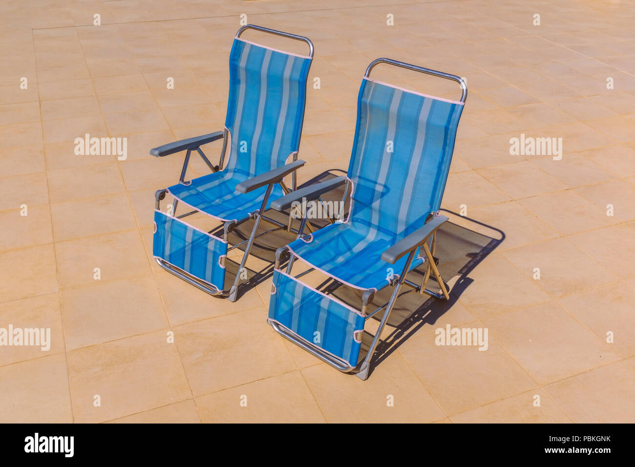Two blue nylon stripe foldable  recliner deck chairs in an upright position with no body sitting on them. They are on a stone patio in bright sunshine Stock Photo