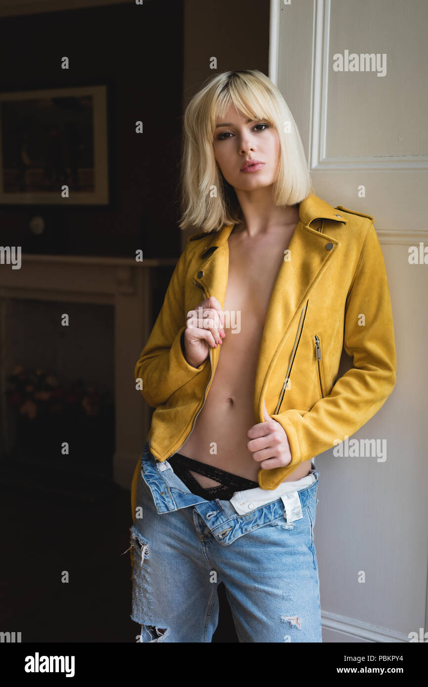 Blonde female standing beside a shuttered window wearing a mustard yellow leather jacket. Her hands are holding the open jacket. Stock Photo