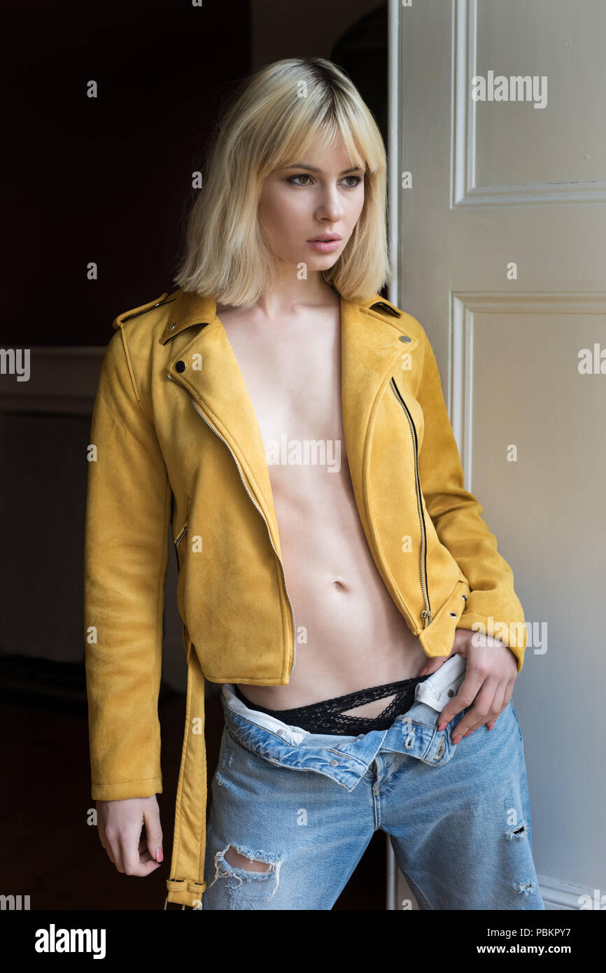 Blonde female standing beside a shuttered window wearing a mustard yellow leather jacket. Her thumb is tucked in her ripped blue jeans waistband. Stock Photo