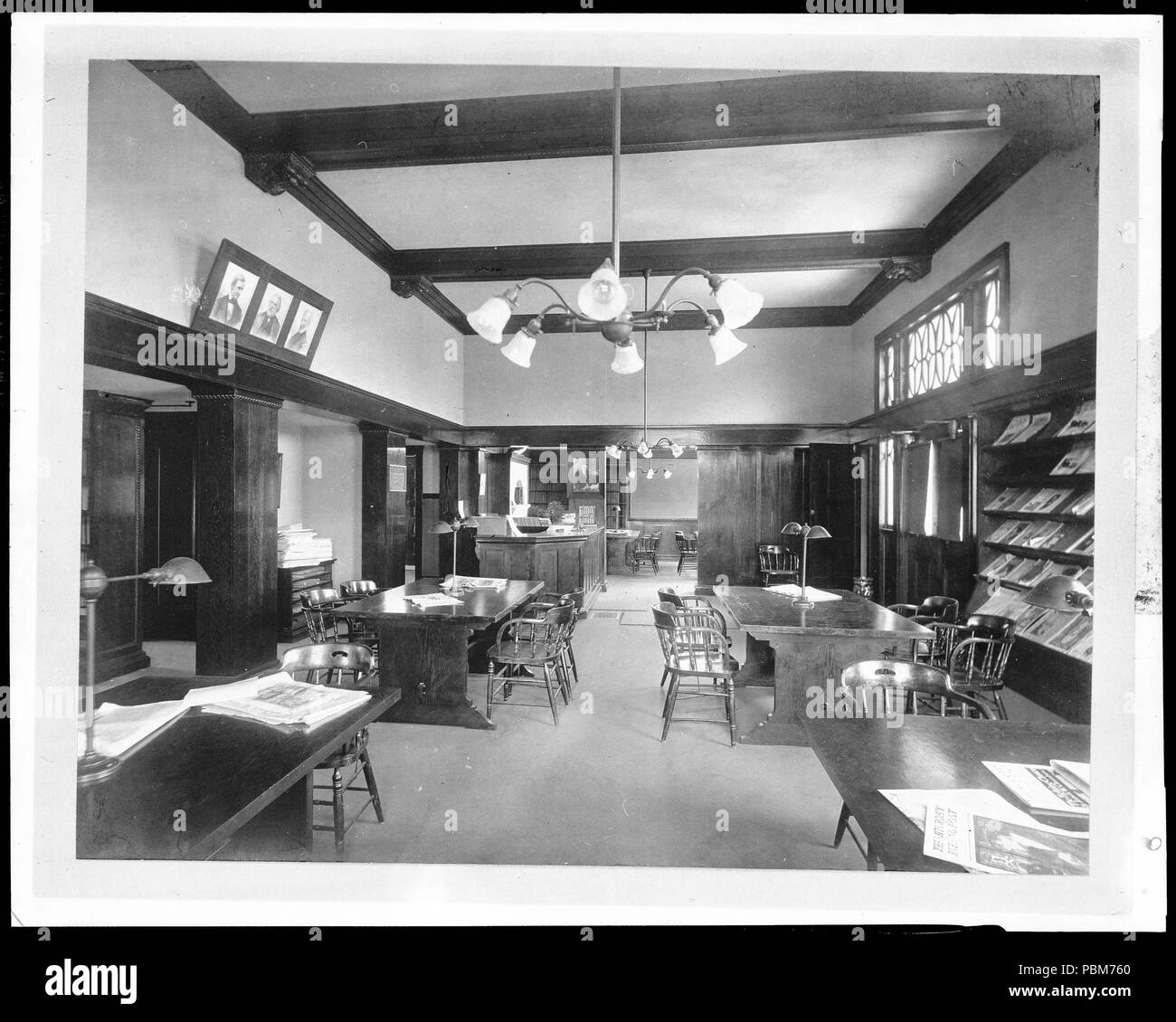 . English: Interior of the Carnegie Public Library in Covina, ca.1908 (1920?) Photograph of the interior of the Carnegie Public Library in Covina, ca.1908 (1920?). Four large wooden tables are arranged in a long rectangular room. A wooden counter is in the back left corner of the room, and a large window is at right. The walls are covered with wooden paneling except on the top third, where they are painted in a light color. There are three portraits hanging on the wall above the paneling at left. Magazine racks line the wall at left, and two chandeliers with six lights each are hanging from th Stock Photo