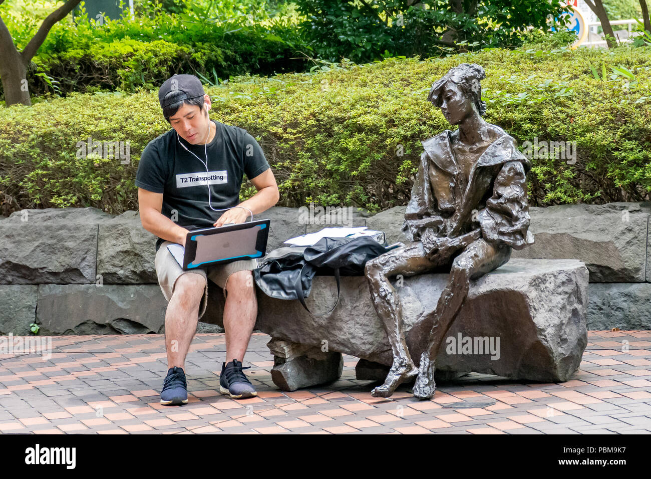 Bronze statue of Japanese lady looks over the shoulder of a young Japanese man working on his laptop, wearing baseball cap and white earphones Stock Photo