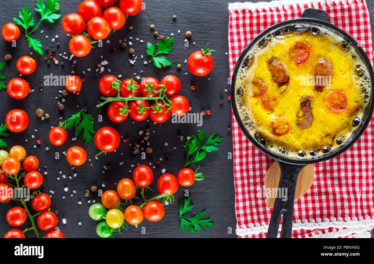 fried omelette with sausages and chicken eggs in a round cast-iron pot and ripe red cherry tomatoes on a black background, top view Stock Photo