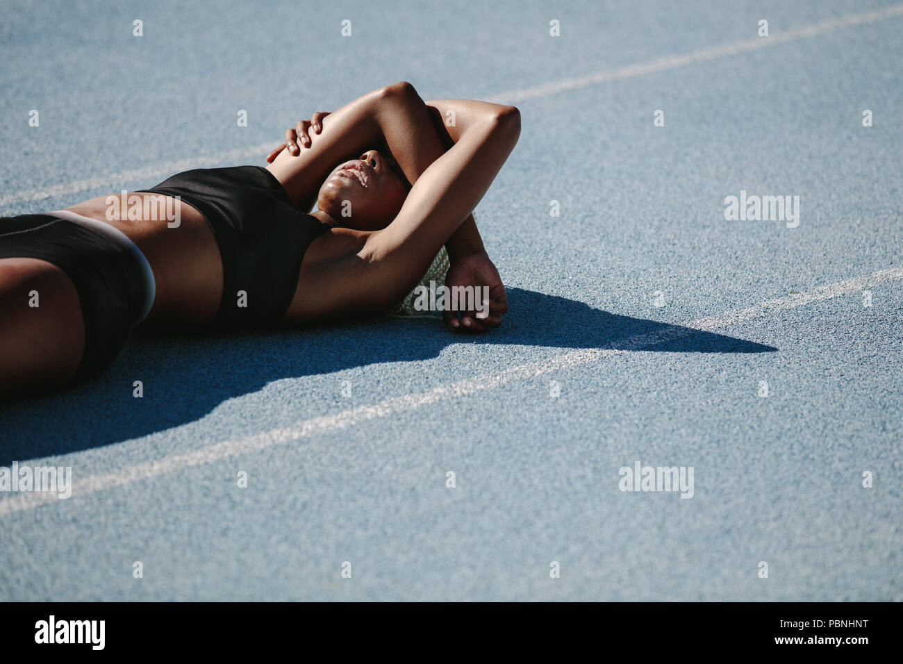 Woman athlete lying on the running track relaxing after workout. Female sprinter resting after workout lying on track on a sunny day. Stock Photo