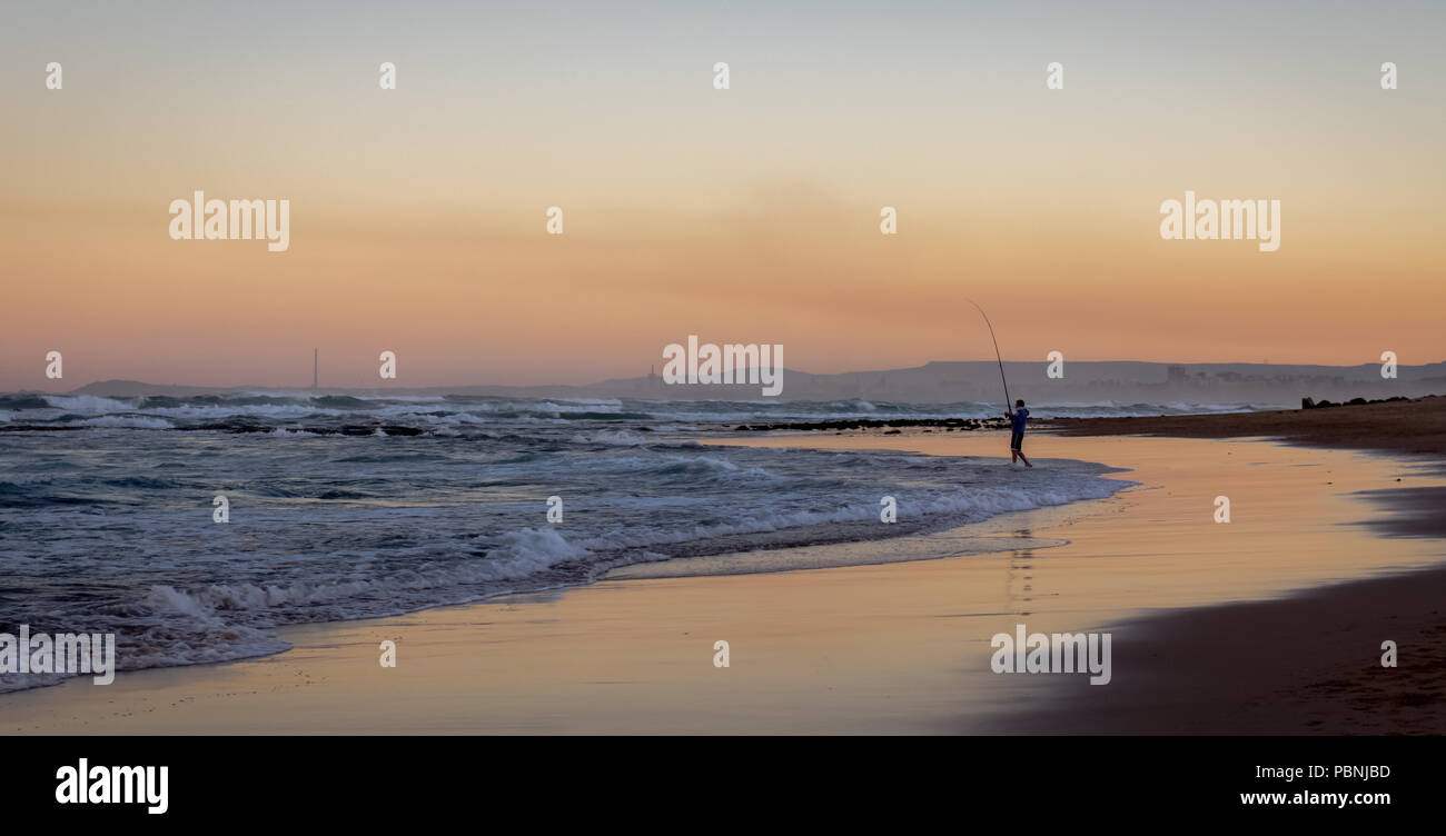 A landscape view at dusk of a silhouette of a distant fisherman fishing off a deserted Australian beach, Australia Stock Photo