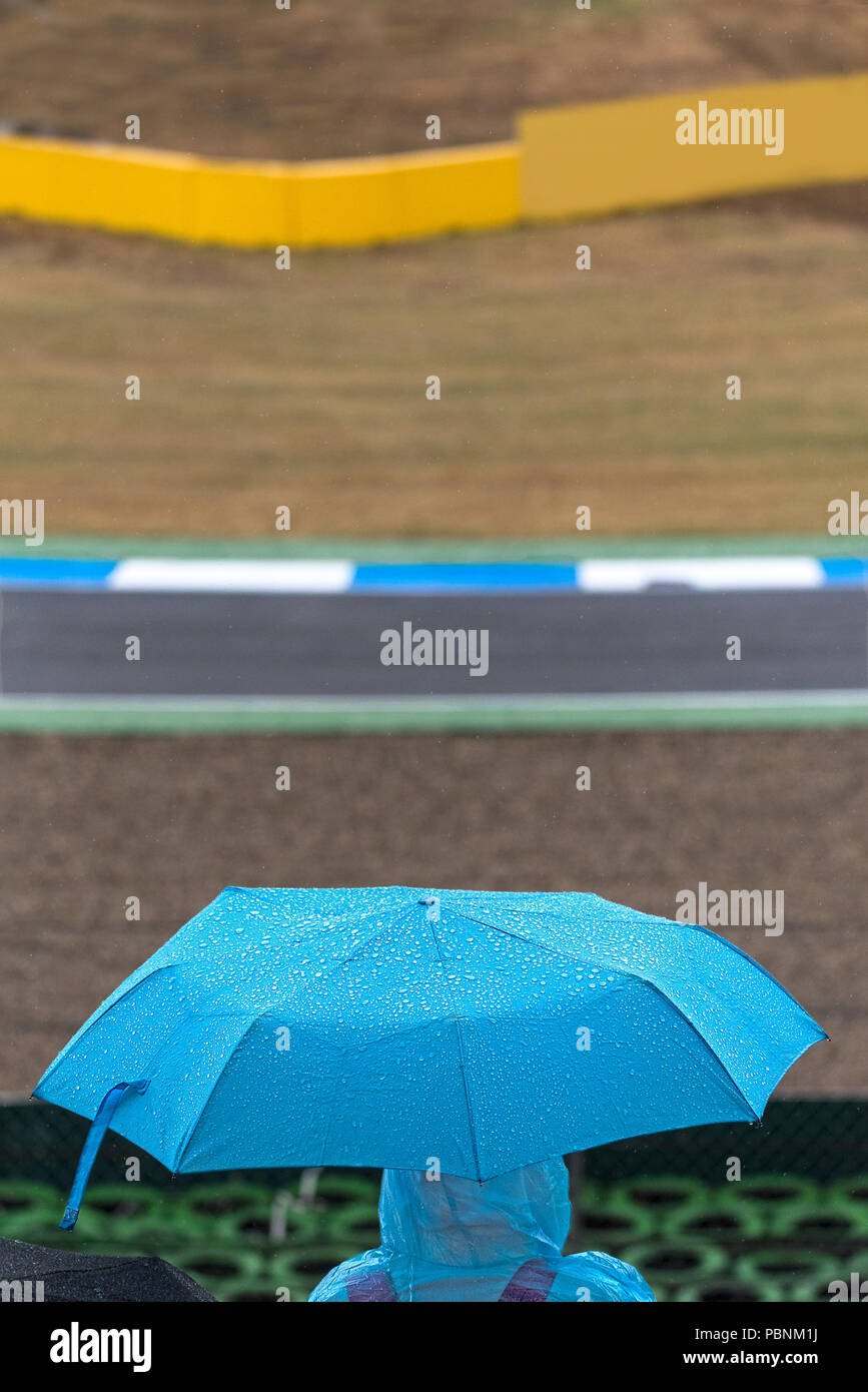 A motorsport spectator shelters from the rain under a blue umbrella and poncho in the grandstands between races. Stock Photo