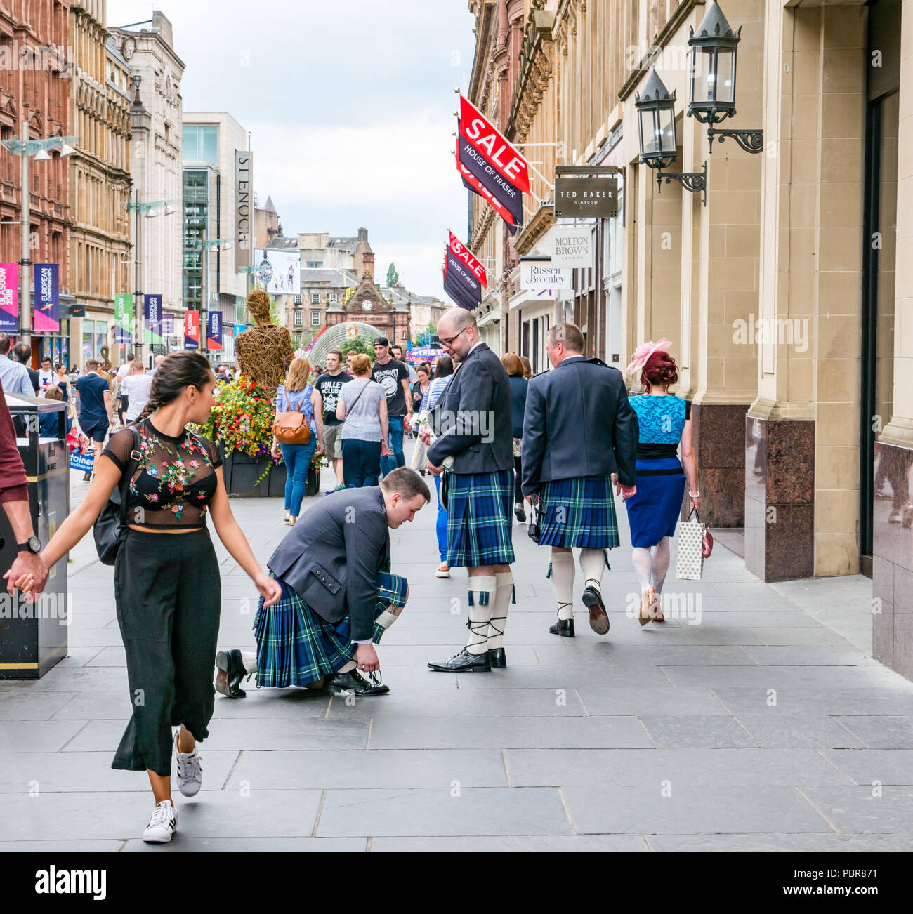 People dressed for wedding with men in kilts stopping to tie shoelace and young woman turning head, Buchanan Street, Glasgow, Scotland, UK Stock Photo