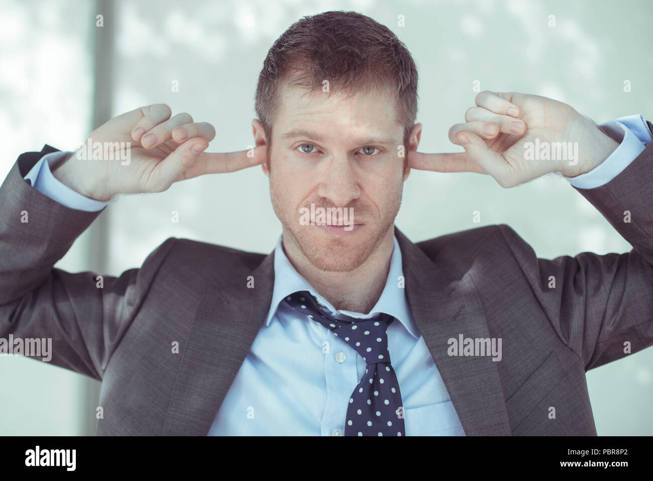 Caucasian male in his 30's wearing a grey suit, unbuttoned shirt and loosened tie,with a serious facial expression, covering his ears with two fingers. Stock Photo