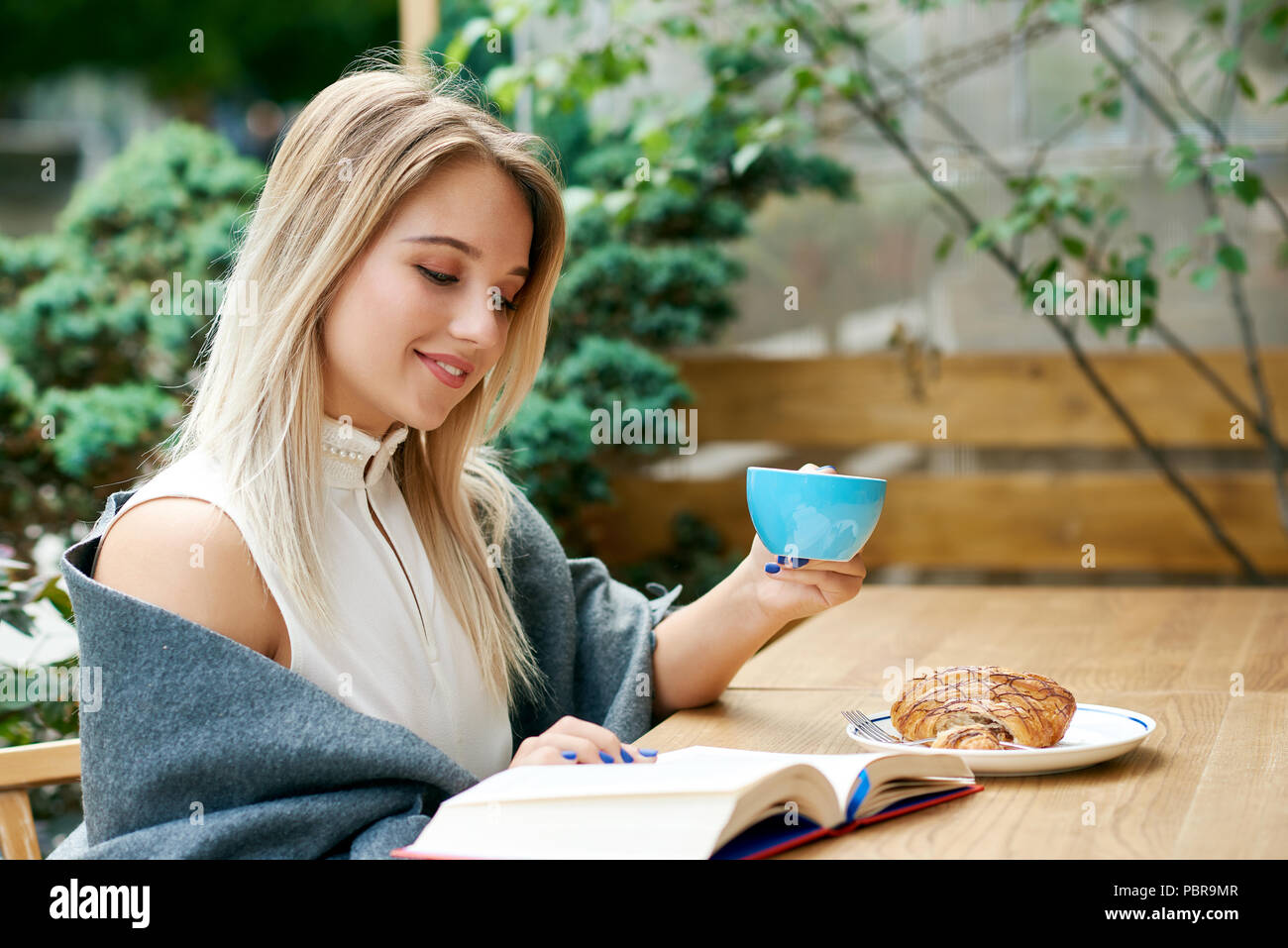 Blonde girl reading a book while drinking coffee on outdoors cafe's lounge. Wearing white blouse decorated with pearls, shoulders covered with grey warm blanket. Smiling, having good time. Stock Photo