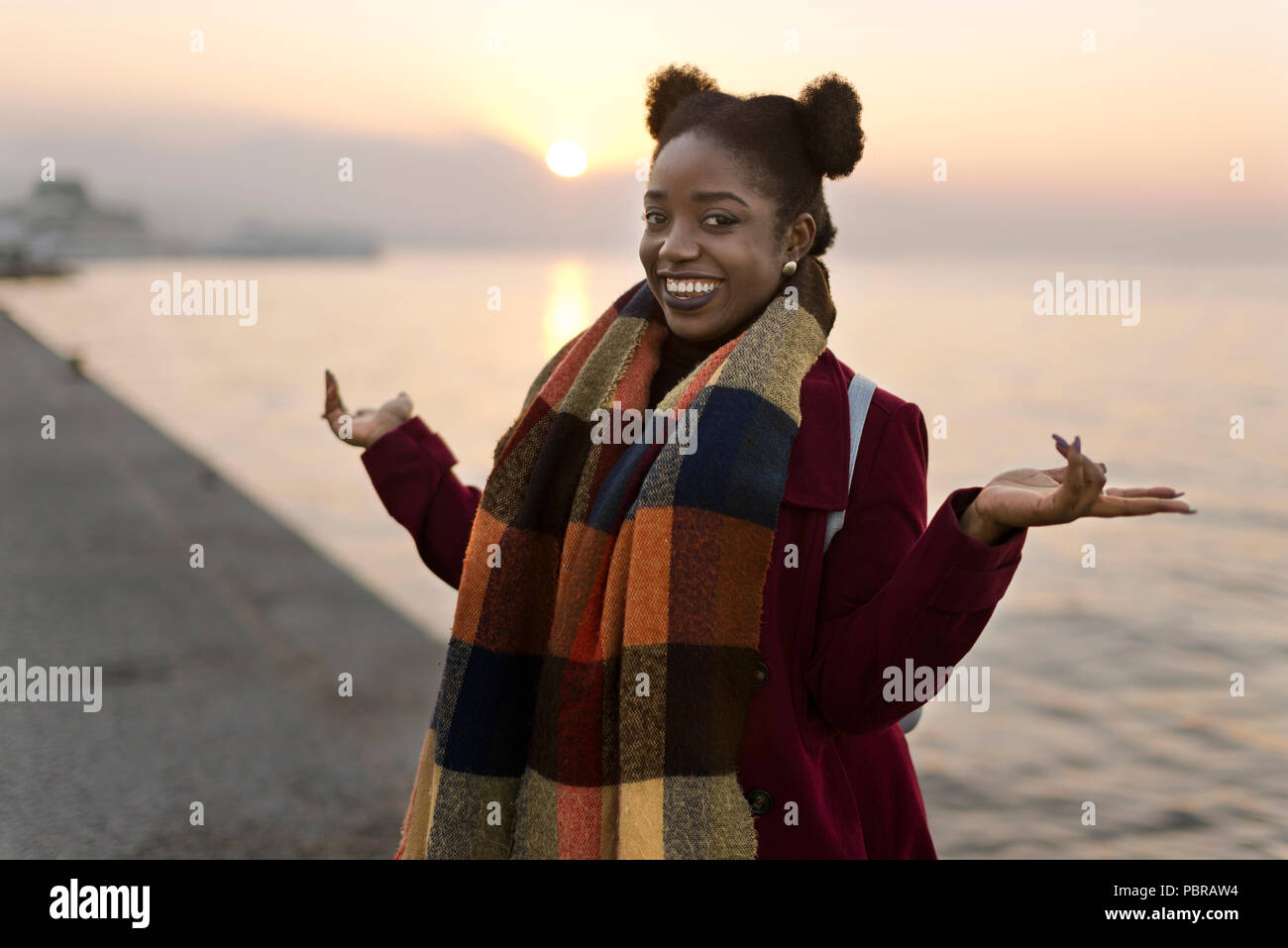 Portrait of a 25 years old African woman with a red coat and weft near to the waters edge with a sunset. Stock Photo